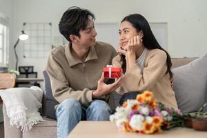 romantisch jung asiatisch Paar Umarmen geben Geschenk im Leben Zimmer beim heim. fallen im Liebe. Valentinstag Konzept foto