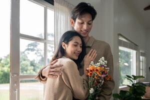 romantisch jung asiatisch Paar Umarmen mit halten Blumen und lächelnd im Leben Zimmer beim heim. fallen im Liebe. Valentinstag Konzept foto