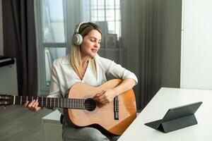 heiter jung Frau im kabellos Kopfhörer mit akustisch Musical Instrument während Sitzung mit Laptop foto