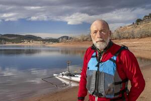 Umwelt Porträt von ein Senior Mann tragen Trockenanzug und Leben Jacke mit ein Rudern Schale auf See Ufer foto