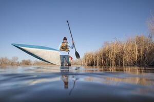 Senior männlich Paddler ist Starten ein Stand oben Paddel Boot auf ein Ruhe See im früh Frühling, Frosch Perspektive von ein Aktion Kamera beim Wasser Niveau foto
