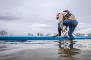 Senior männlich ist Anbringen ein Sicherheit Leine Vor Paddeln ein Stand oben Paddel Boot auf ein See im früh Frühling, Frosch Perspektive von ein Aktion Kamera beim Wasser Niveau foto