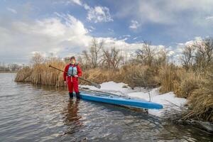 Senior Paddler tragen Leben Jacke und Trockenanzug ist Stehen im Wasser Nächster zu seine Stand oben Paddel Boot auf See im Colorado im Winter oder früh Frühling Landschaft foto