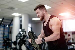 Porträt von gut aussehend muskulös Powerlifter mit Gewicht Platte im seine Hände im das Sport Fitnessstudio. Bodybuilder mit schwer Platte Züge seine Bizeps mit das öffnen Mund. Nahansicht foto