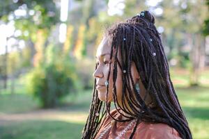 Porträt von schön europäisch Frau mit lange afrikanisch Zöpfe und ethnisch Dekorationen draußen im ein Park. foto