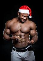 Porträt von ein stark Afroamerikaner Mann tragen Weihnachten Hut zeigen aus seine Körperbau gegen zurück Hintergrund. Mann mit perfekt Körper posieren beim das Studio während suchen beim das Kette foto