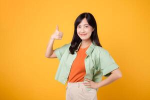 fesselnd jung asiatisch Frau im ihr 30er Jahre, gekleidet im ein Orange Hemd und Grün Jumper. ihr Daumen oben Geste, isoliert auf ein sonnig Gelb Hintergrund, verkörpert das Konzept von Unterstützung. foto
