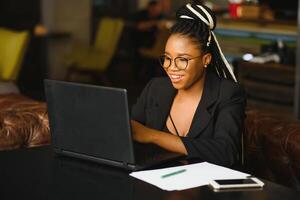 jung Mädchen im Brille erstaunlich suchen im Laptop beim Cafe. afrikanisch amerikanisch Mädchen Sitzung im Restaurant mit Laptop und Tasse auf Tisch. Porträt von überrascht Dame mit dunkel lockig Haar im Kopfhörer foto