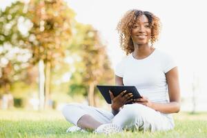 nachdenklich süß gemischt weiblich International Schüler mit lockig Haar ist Sitzung auf frisch Gras mit modern Laptop im Öffentlichkeit Park, gelehnt auf Apfel Baum und wehmütig suchen beiseite während ihr brechen foto