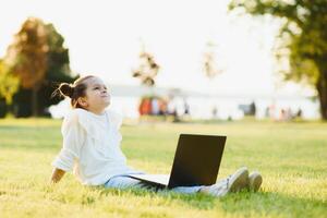 wenig Mädchen Sitzung im das Park und Arbeiten mit Laptop. Ausbildung, Lebensstil, Technologie Konzept foto