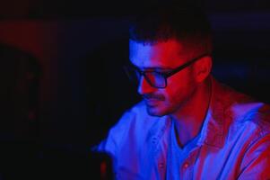 Profi Spieler. tausendjährig Kerl mit Desktop Computer spielen online Videospiel Sitzung beim Nacht beim heim. Neon- Beleuchtung, selektiv Fokus foto