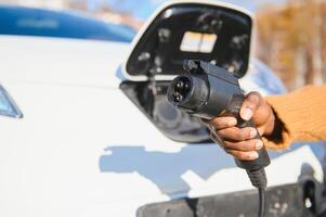 schließen oben von männlich Hand halten elektrisch Laden Stecker zum Luxus rot Wagen. afrikanisch Geschäftsmann Laden seine Auto auf Bahnhof. Öko freundlich Transport. foto