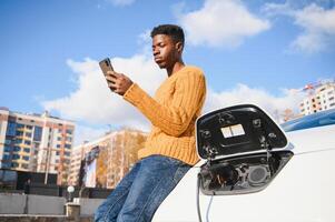 urban, elektrisch Fahrzeug, Öko Konzept. jung schwarz gehäutet Mann, warten zum seine elektrisch Auto Laden foto