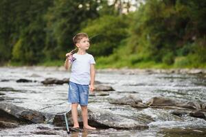 süß Junge im Weiß t Hemd Angeln im das Fluss und hat Spaß, lächelt. Ferien mit Kinder, Feiertage, aktiv Wochenenden Konzept foto