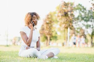 schön dunkel gehäutet Hipster Mädchen reden auf Handy, Mobiltelefon Telefon Sitzung auf Grün Gras neu erstellen draußen, afrikanisch amerikanisch Frau tausendjährig Sitzung auf Grün Gras reden auf zellular und gut Tarife foto