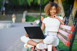 schön jung gemischt Rennen Frau Arbeiten mit Laptop und mit Telefon. Sitzung auf das Bank im Park, draussen Porträt , trägt beiläufig Kleider und Brille foto