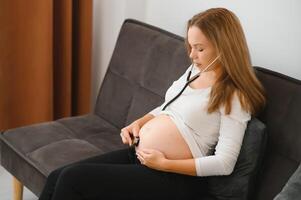 schwanger Frau mit Stethoskop Prüfung ihr Bauch auf Sofa, glücklich schwanger Frau Konzept. foto