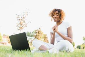 nachdenklich süß gemischt weiblich International Schüler mit lockig Haar ist Sitzung auf frisch Gras mit modern Laptop im Öffentlichkeit Park, gelehnt auf Apfel Baum und wehmütig suchen beiseite während ihr brechen foto