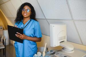 jung afro amerikanisch weiblich Arzt Stehen im Klinik Büro in der Nähe von modern Ultraschall Maschine. foto