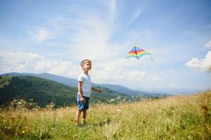 wenig Junge Laufen auf ein Hintergrund von Berge mit Drachen. sonnig Sommer- Tag. glücklich Kindheit Konzept. foto