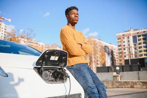 schließen oben von afrikanisch amerikanisch Mann verbinden Laden Kabel zu elektrisch Wagen. jung männlich Stehen in der Nähe von seine modern Auto mit Leder Koffer im Hand. foto