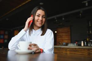 Junge charmante Frau, die mit dem Handy anruft, während sie in der Freizeit allein im Café sitzt, attraktive Frau mit süßem Lächeln, die sich mit dem Handy unterhält, während sie sich im Café ausruht foto