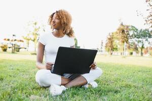 nachdenklich süß gemischt weiblich International Schüler mit lockig Haar ist Sitzung auf frisch Gras mit modern Laptop im Öffentlichkeit Park, gelehnt auf Apfel Baum und wehmütig suchen beiseite während ihr brechen foto