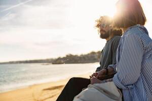 schön romantisch Paar Sitzung beim das Strand tragen beiläufig Kleider und suchen beim das Ozean foto