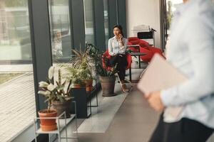 Frau Büro Arbeiter Sitzung in der Nähe von Fenster und reden durch Telefon mit Kollege auf Hintergrund foto