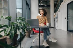 lächelnd weiblich Analytiker im Brille Arbeiten auf Laptop und Herstellung Anmerkungen während Sitzung im gemütlich Büro foto