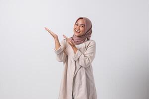 Porträt von aufgeregt asiatisch Hijab Frau im beiläufig passen zeigen und zeigen Produkt im ihr Seite mit Finger. Geschäftsfrau Konzept. isoliert Bild auf Weiß Hintergrund foto