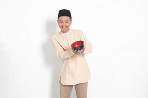 Porträt von aufgeregt asiatisch Muslim Mann im koko Hemd mit Schädeldecke zeigen und halten ein leeren Platte. Schüssel Vorlage zum Essen Marke. isoliert auf Weiß Hintergrund foto