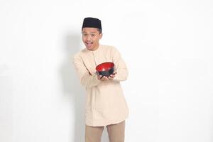 Porträt von aufgeregt asiatisch Muslim Mann im koko Hemd mit Schädeldecke zeigen und halten ein leeren Platte. Schüssel Vorlage zum Essen Marke. isoliert auf Weiß Hintergrund foto