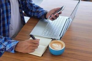 schließen oben von männlich Hand Schreiben auf Hinweis Buch mit Stift, zeigen leer Bildschirm von Laptop und Smartphone. Arbeiten im Cafe Konzept mit ein Tasse von Kaffee foto