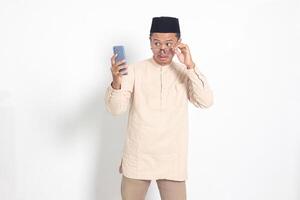 Porträt von überrascht asiatisch Muslim Mann im koko Hemd mit Schädeldecke halten Handy, Mobiltelefon Telefon, zeigen Beeindruckend schockiert Gesicht Ausdruck. Werbung und Sozial Medien Konzept. isoliert Bild auf Weiß Hintergrund foto