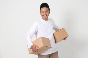 Porträt von glücklich asiatisch Muslim Mann im koko Hemd mit peci suchen zuversichtlich während Tragen Karton Kasten. Heimkehr auf eid Mubarak Konzept. isoliert Bild auf Weiß Hintergrund foto