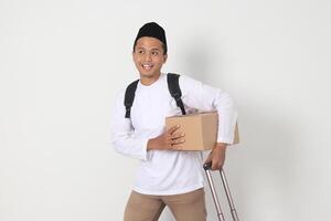 Porträt von aufgeregt asiatisch Muslim Mann im koko Hemd mit peci Tragen Karton Box und halten Koffer handhaben. gehen Zuhause zum eid Mubarak. isoliert Bild auf Weiß Hintergrund foto