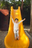 froh Mädchen gleiten Nieder Gelb Spielplatz gleiten. aufgeregt jung Mädchen mit Waffen angehoben hoch gleiten Nieder ein hell Gelb rutschen beim ein Spielplatz. Aktivität zum Kind Spielplatz Konzept. foto