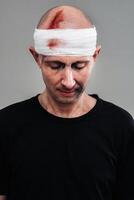 gegen ein grau Hintergrund steht ein angeschlagen und angeschlagen Mann im ein schwarz T-Shirt mit seine Kopf eingewickelt im ein Binde foto
