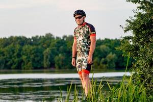 Porträt von ein gut aussehend männlich Radfahrer Stehen in der Nähe von Fluss gegen ein schön Landschaft mit ein See. Mann suchen weg. foto