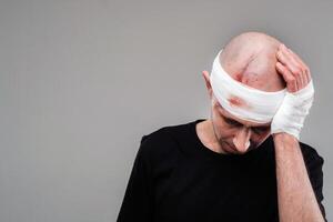 ein angeschlagen und angeschlagen Mann im ein schwarz T-Shirt steht gegen ein grau Hintergrund, halten seine Schmerz Kopf mit seine Hände eingewickelt um es foto
