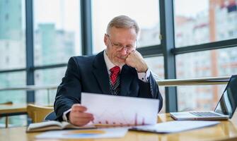 nachdenklich Mitte alt Geschäftsmann im passen mit ein Laptop auf Tabelle während Arbeiten mit Unterlagen. foto