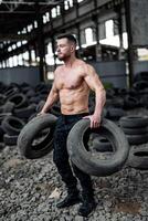 stark jung Mann Aufzüge Reifen mit nacktem Oberkörper. groß Muskeln und perfekt Abs. viele Reifen auf das Hintergrund. starker Mann hat trainieren foto