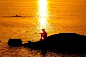 Sonnenuntergang Silhouette von Sitzung Mann auf ein See. draussen Orange Sonnenlicht von Mann haben ein ausruhen. foto