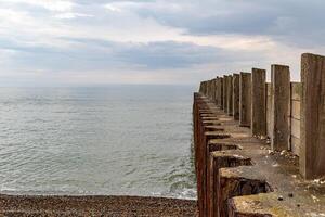 Küsten Verteidigung hölzern Buhnen auf Kieselstein Strand führen in Ruhe Meer unter wolkig Himmel. foto