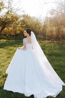 Brünette Braut im ein Weiß Kleid mit ein lange Zug, halten ein Strauß und Gehen im das Garten. Hochzeit Foto Session im Natur. schön Haar und bilden. Feier