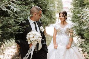 ein Hochzeit Paar ist Gehen im Natur auf ein Herbst Tag. glücklich jung Braut und elegant Bräutigam halten Hände. ein stilvoll Paar von Jungvermählten auf ihr Hochzeit Tag. foto