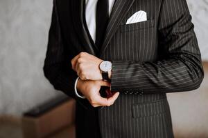 ein Nahansicht von ein abgeschnitten Rahmen von ein Mann setzt auf ein Uhr mit ein Leder Gürtel, ist gekleidet im ein stilvoll Anzug, ein Weiß Shirt, trägt ein Gold Ring. foto