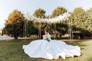 ein Brünette Braut im ein Schleier und ein Tiara im ihr Haar sitzt auf ein Verbreitung Kleid, posieren in der Nähe von ein Weiß Bogen foto
