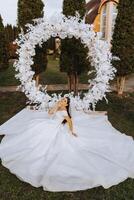ein Brünette Braut im ein Schleier und ein Tiara im ihr Haar sitzt auf ein Verbreitung Kleid, posieren in der Nähe von ein Weiß Bogen gemacht von Blumen im das gestalten von ein Kreis. auf ein Weiß Hintergrund. sonnig Tag. Hochzeit Zeremonie foto
